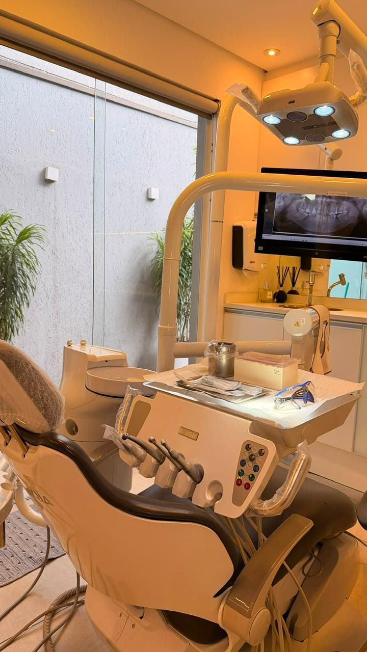 Consultório da Ellos com equipamento marfim, luz âmbar e vidro do chão ao teto para a vegetação