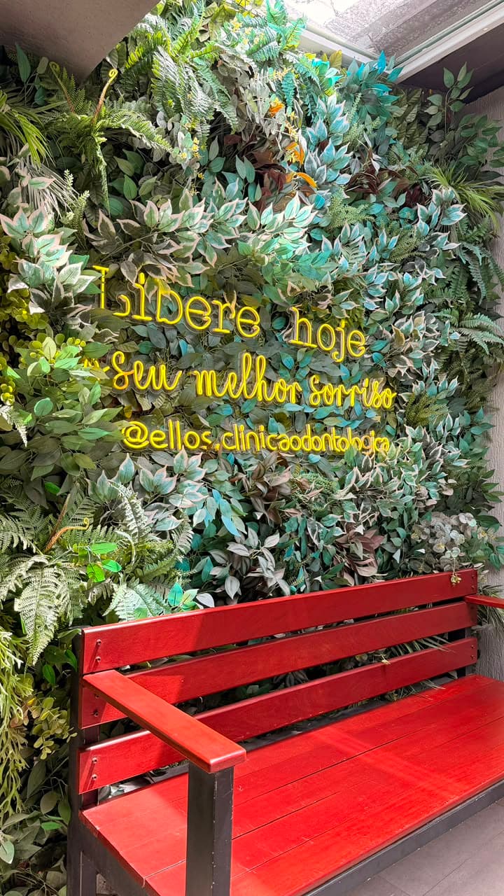 Jardim vertical na entrada da Ellos com neon amarelo manuscrito Libere hoje seu melhor sorriso e banco vermelho de madeira