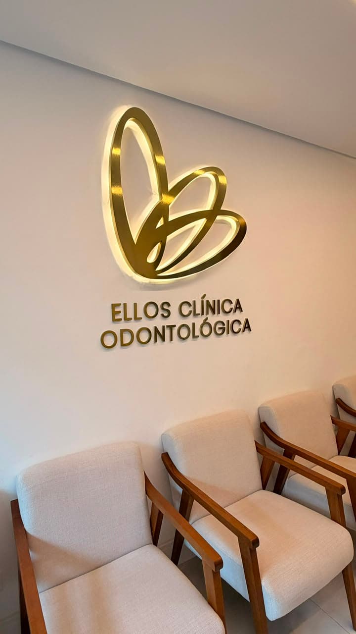 Sala de espera da Ellos Clínica Odontológica com poltronas em linho cru e logo dourado retroiluminado